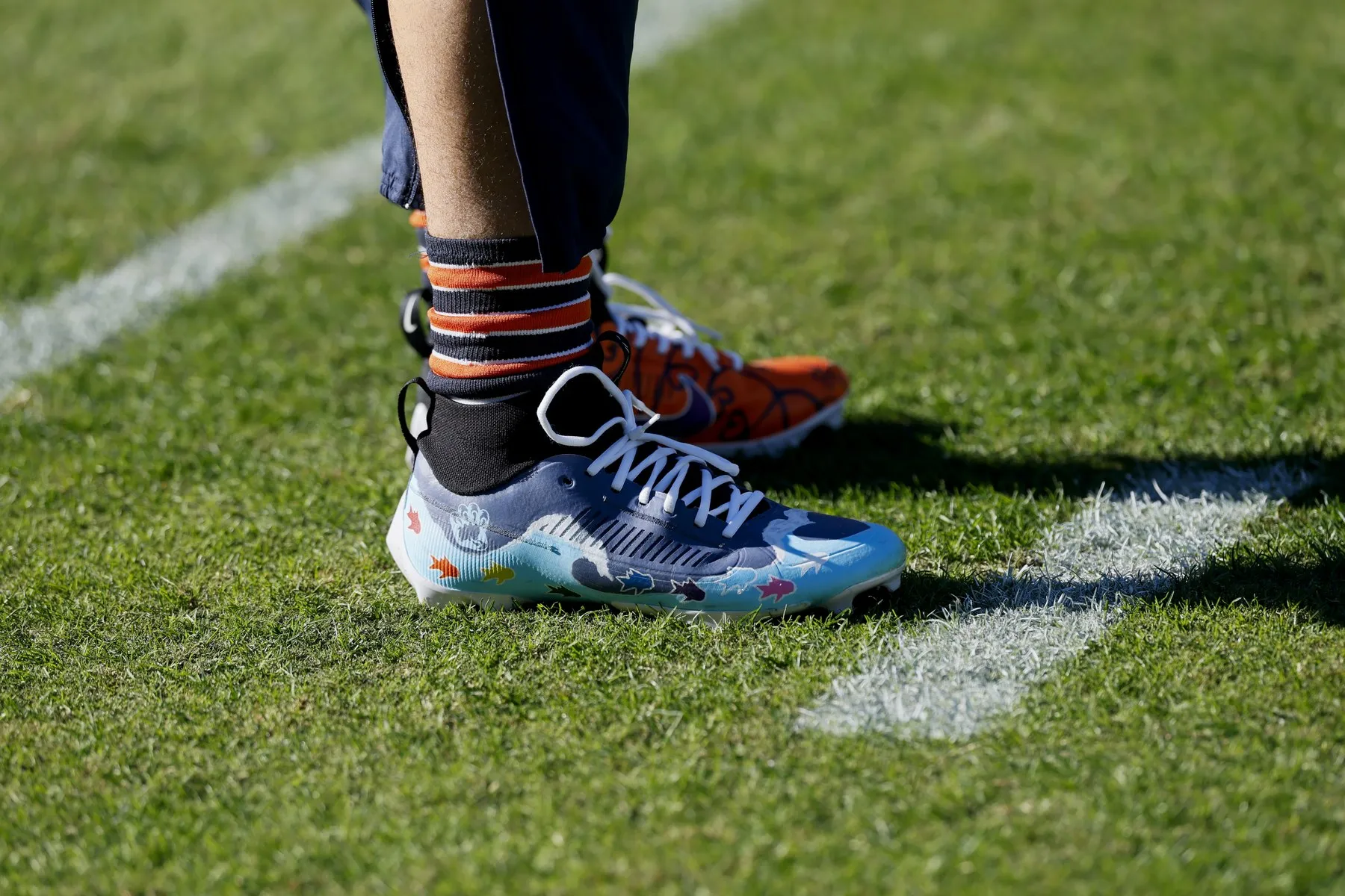 Custom painted cleats worn on a football pitch, one sky blue design on foot and an orange pair beside it on the grass